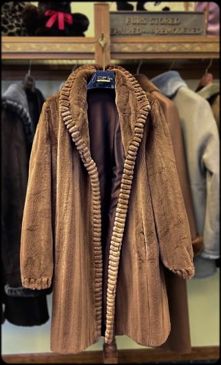 A brown faux fur coat with ribbed trim along the open front, displayed in what appears to be a furrier's shop with other coats and a service sign in the background.