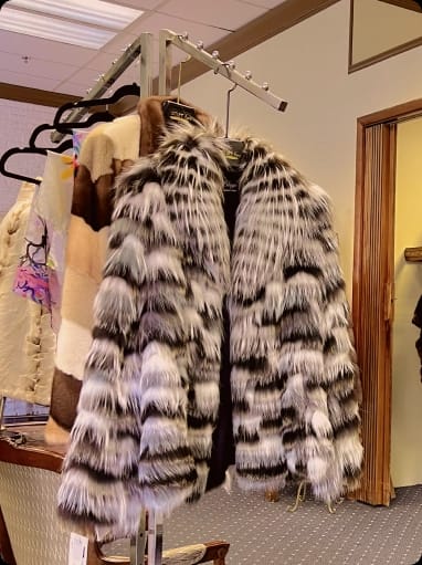 Silver fox fur coat with black and white stripes hanging on a display rack with other fur coats in a store setting.