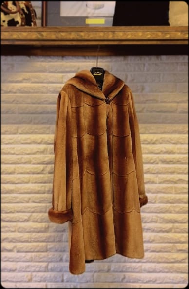 A hooded brown fur coat with wavy horizontal seams and a single button closure hangs on a hanger against a light brick wall.