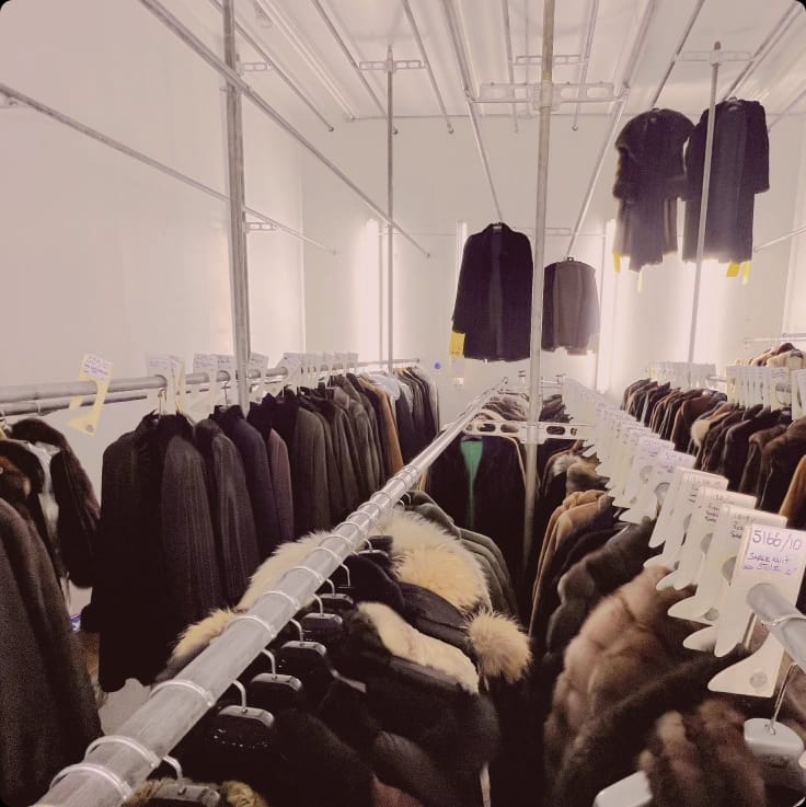Numerous dark fur and winter coats hang on metal racks in a brightly lit storage facility, many with price or inventory tags.