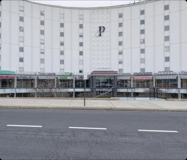 A large, curved white building, "The Pavilion," with a prominent "P" logo and ground-level shops including a post office and pharmacy, viewed from across a street.