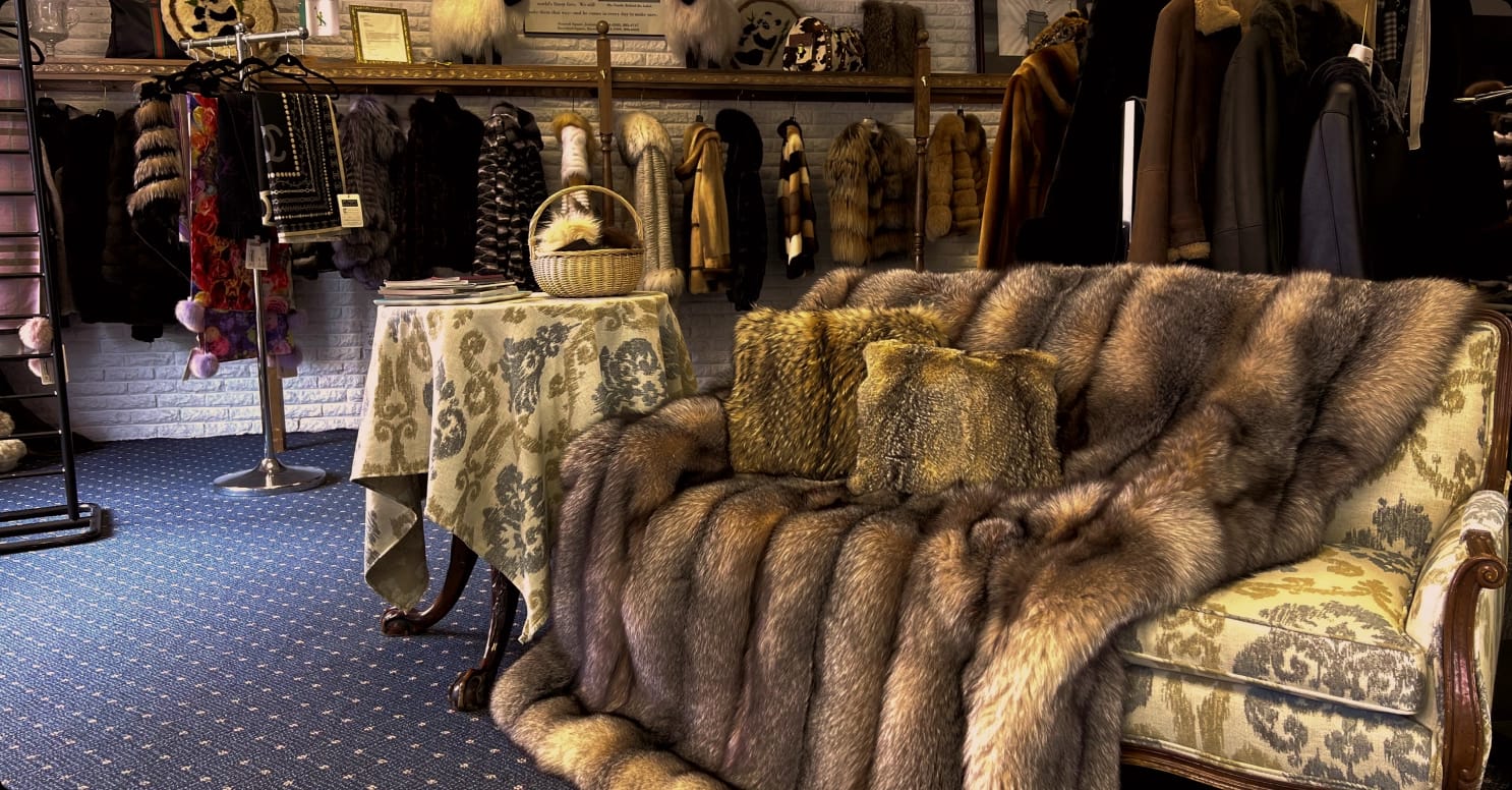 Luxurious showroom featuring a fur-draped sofa with fur pillows, a display of various fur coats and accessories, and a small table with a wicker basket.