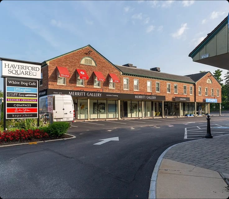 A long, two-story brick building houses Haverford Square shops including Merritt Gallery, J.McLaughlin, and White Dog Cafe, with a parking lot in the foreground under a clear sky.