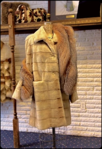 A light brown, horizontally paneled fur coat on a mannequin, accessorized with a reddish-brown fur stole, against a white brick wall with a shelf of bags.