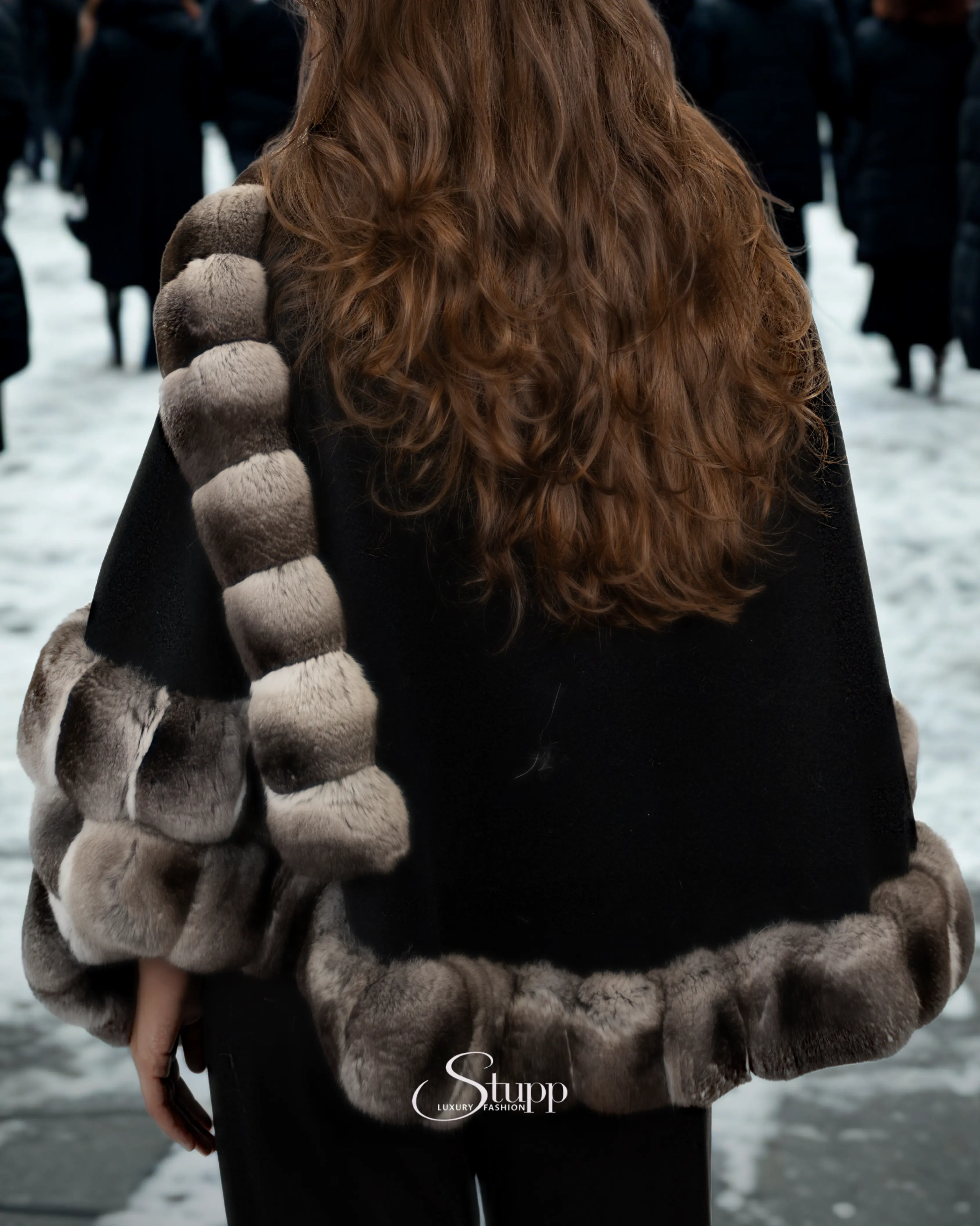 Woman from behind wearing a black cape with luxurious grey chinchilla fur trim and long brown hair, in a snowy outdoor scene. 'Stupp Luxury Fashion' logo at bottom.